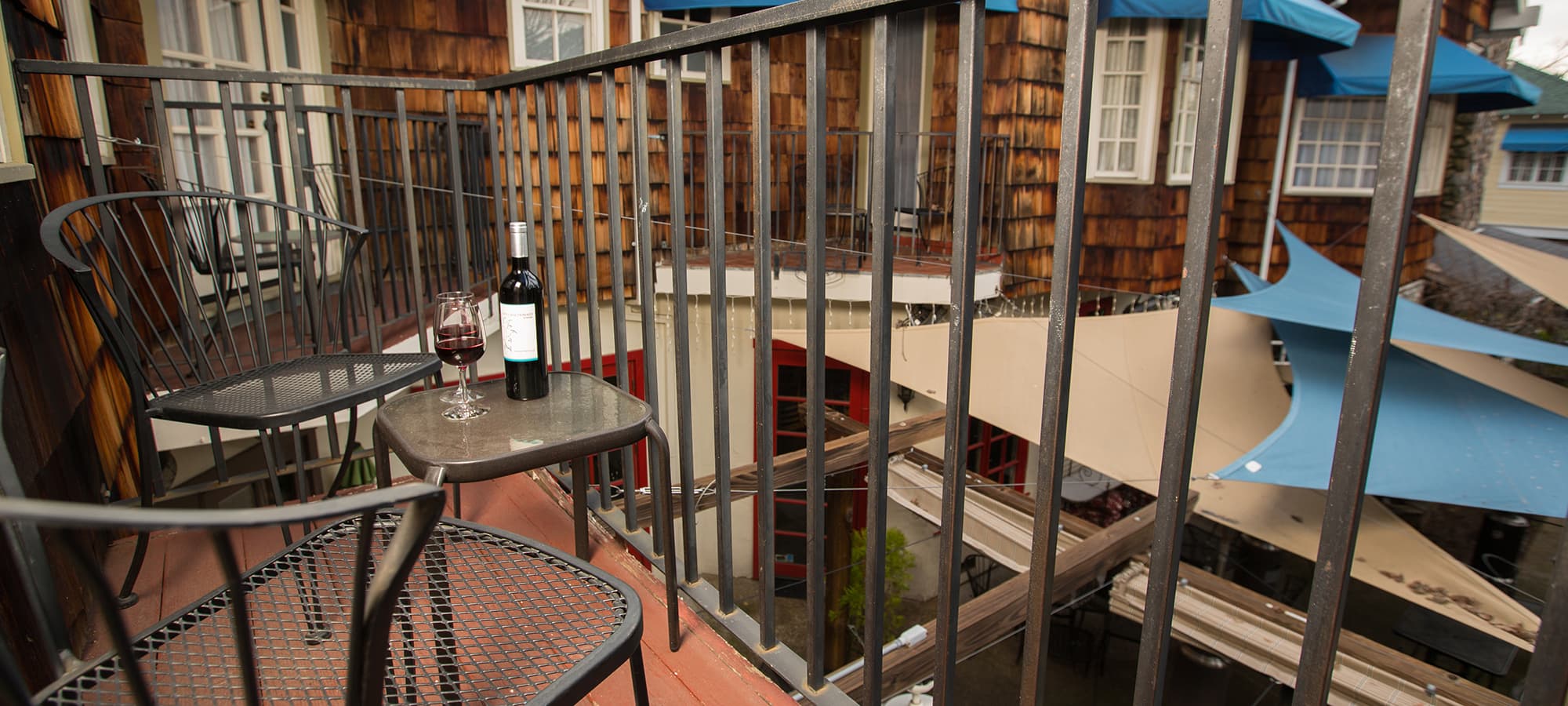 A small balcony with two chairs, a table, a bottle of wine, and glasses overlooking a courtyard.