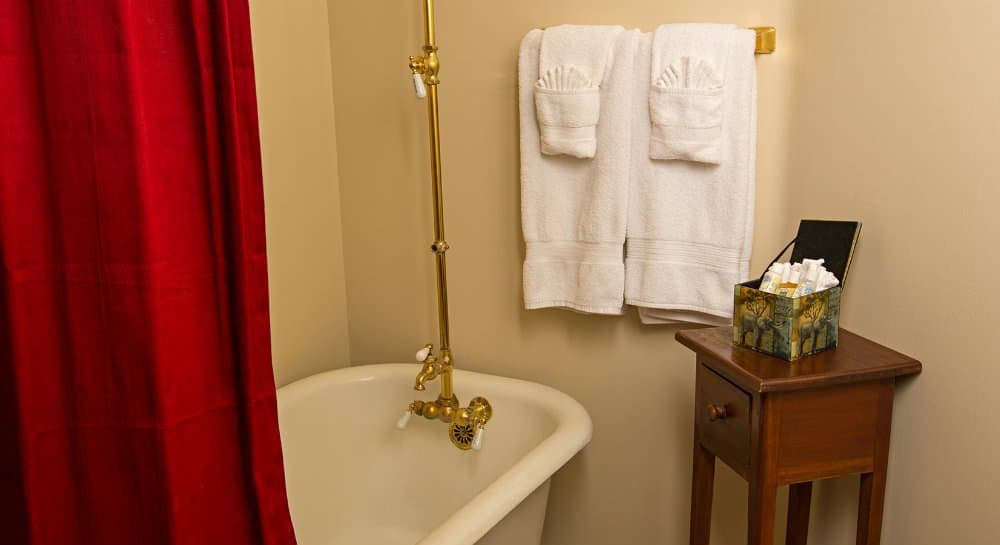 A vintage bathroom featuring a clawfoot tub, red shower curtain, neatly hung towels, and a wooden side table with toiletries.