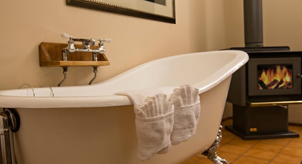 A vintage white bathtub with two towels draped over the edge, beside a cozy wood stove.