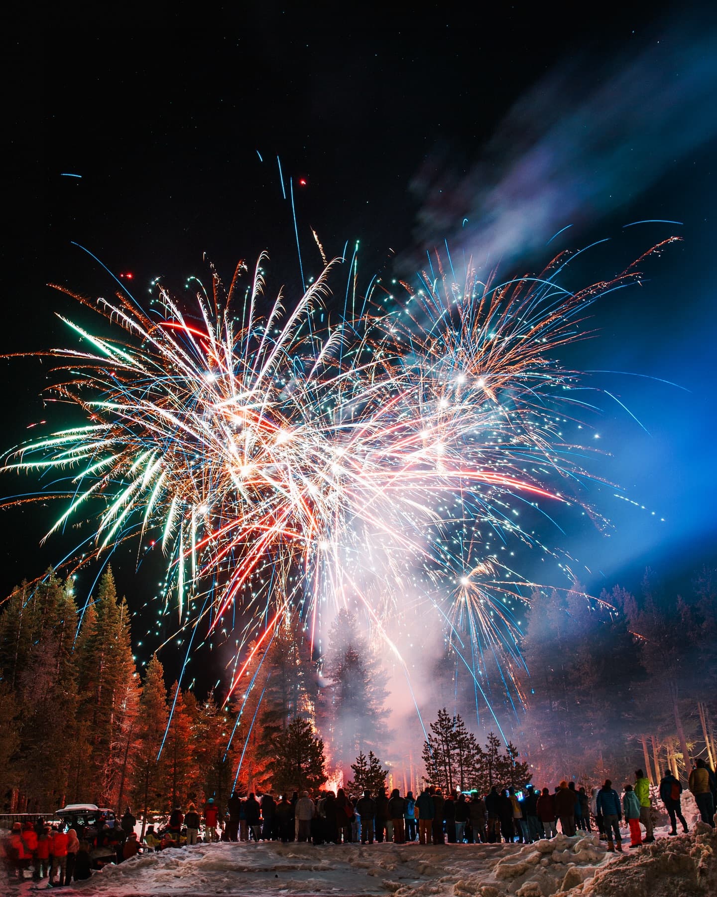 Fireworks illuminate the night sky over a crowd in a snowy landscape.