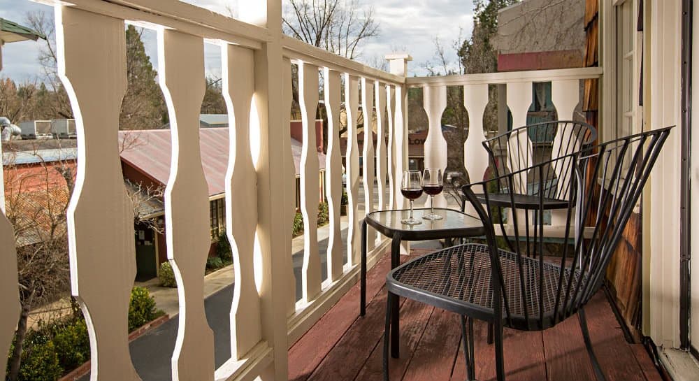 A cozy balcony with two black chairs, a small table, and two glasses of red wine, overlooking a picturesque view.