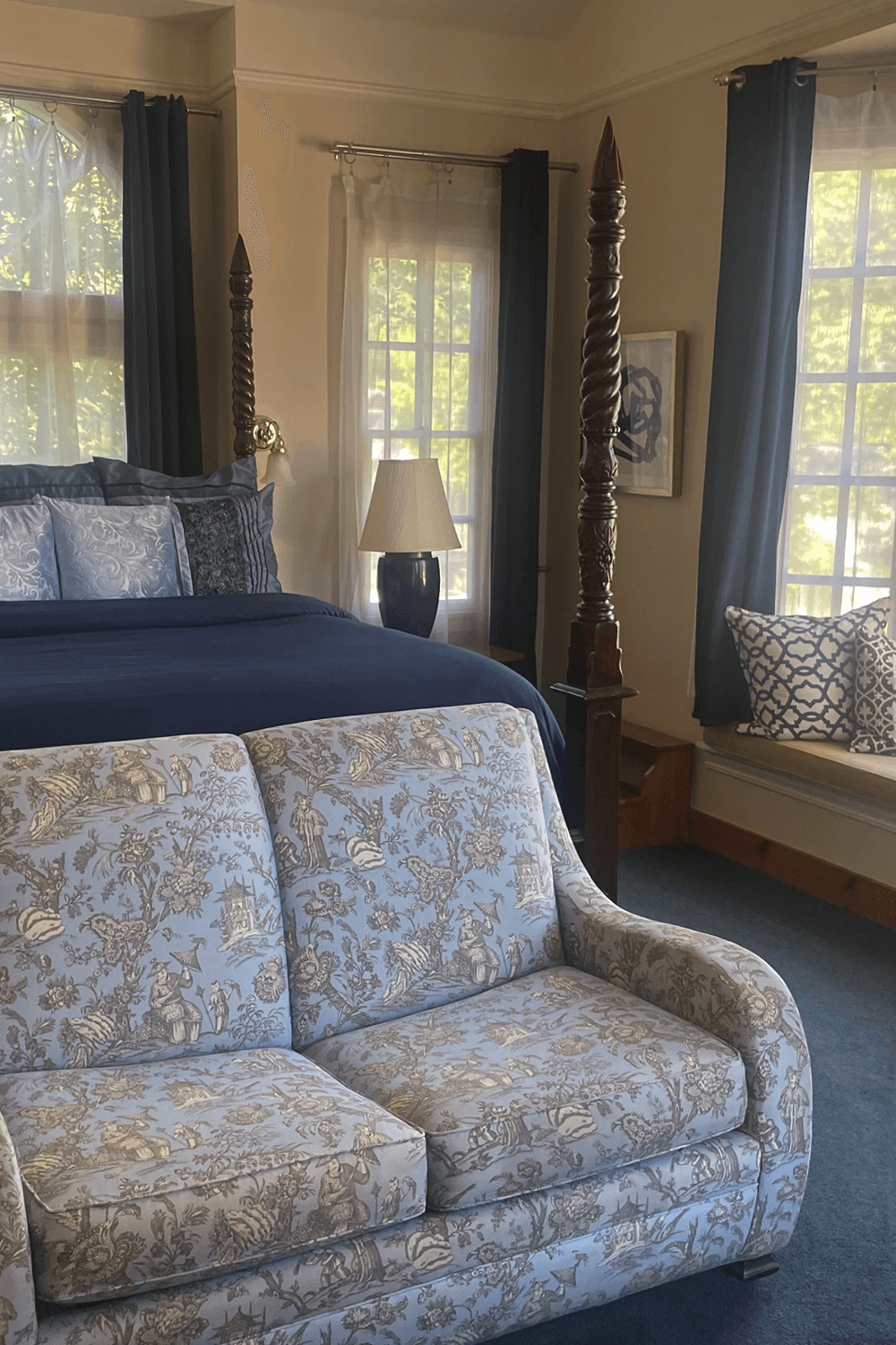 A cozy bedroom featuring a blue floral-patterned sofa in front of a four-poster bed and large windows.