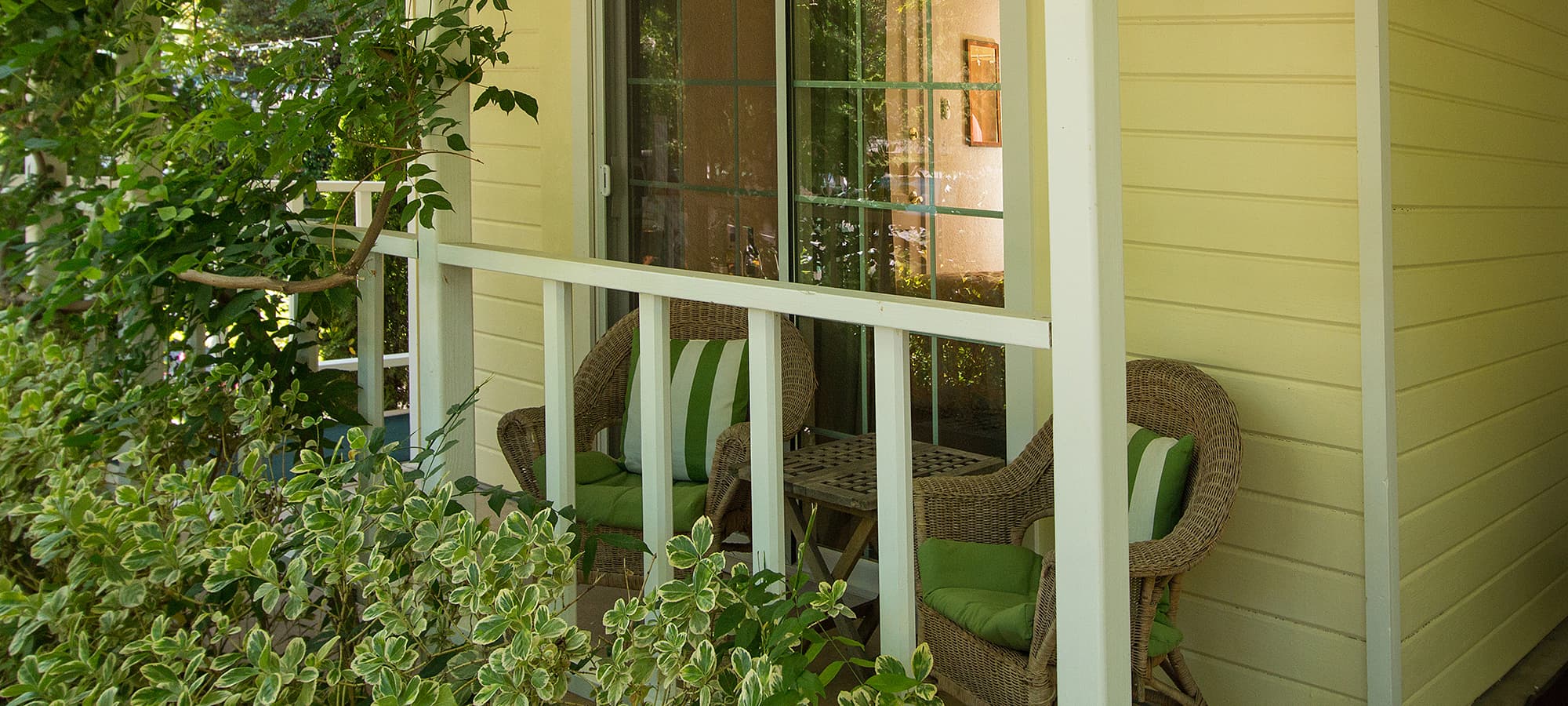 A cozy porch featuring two wicker chairs with green cushions surrounded by lush greenery.