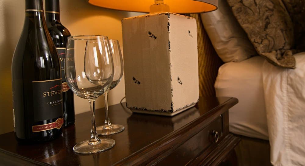 A bedside table featuring two wine bottles, two glasses, and a lamp.