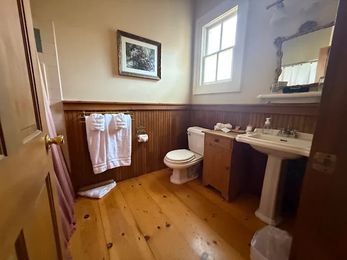 A quaint bathroom featuring wooden accents, a toilet, sink, towels, and natural light from a window.