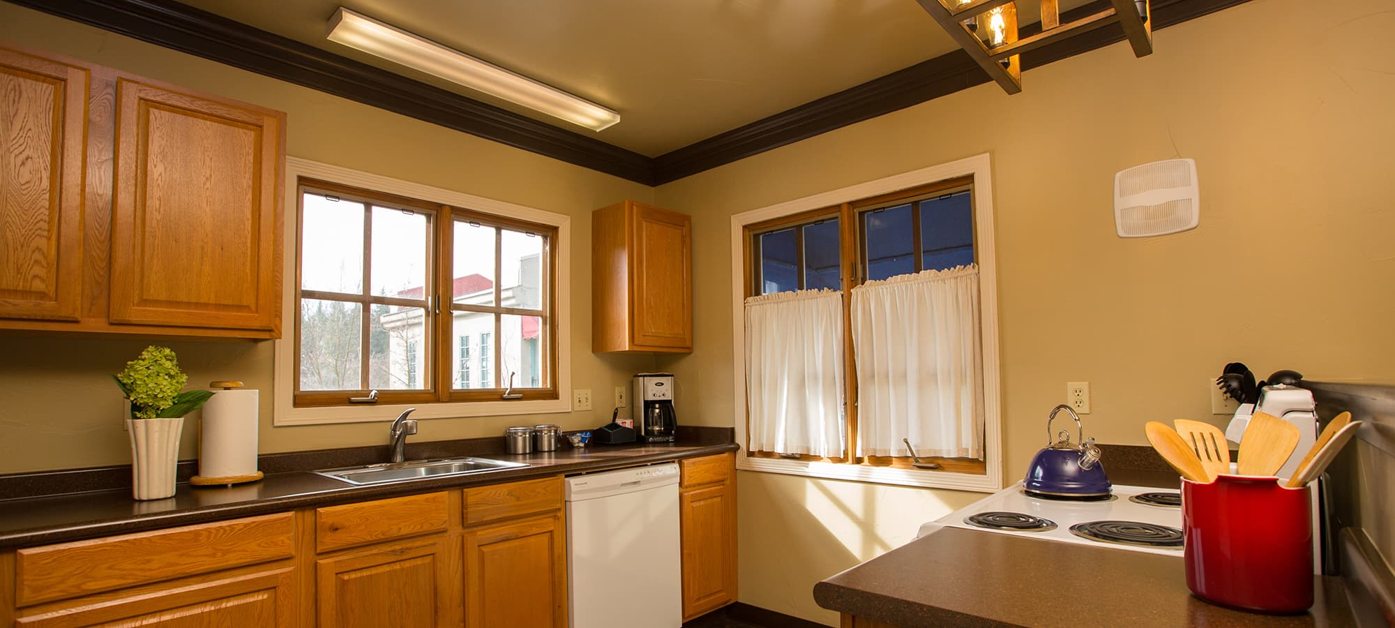 A cozy kitchen featuring wooden cabinetry, a sink, stove, and large windows with curtains.