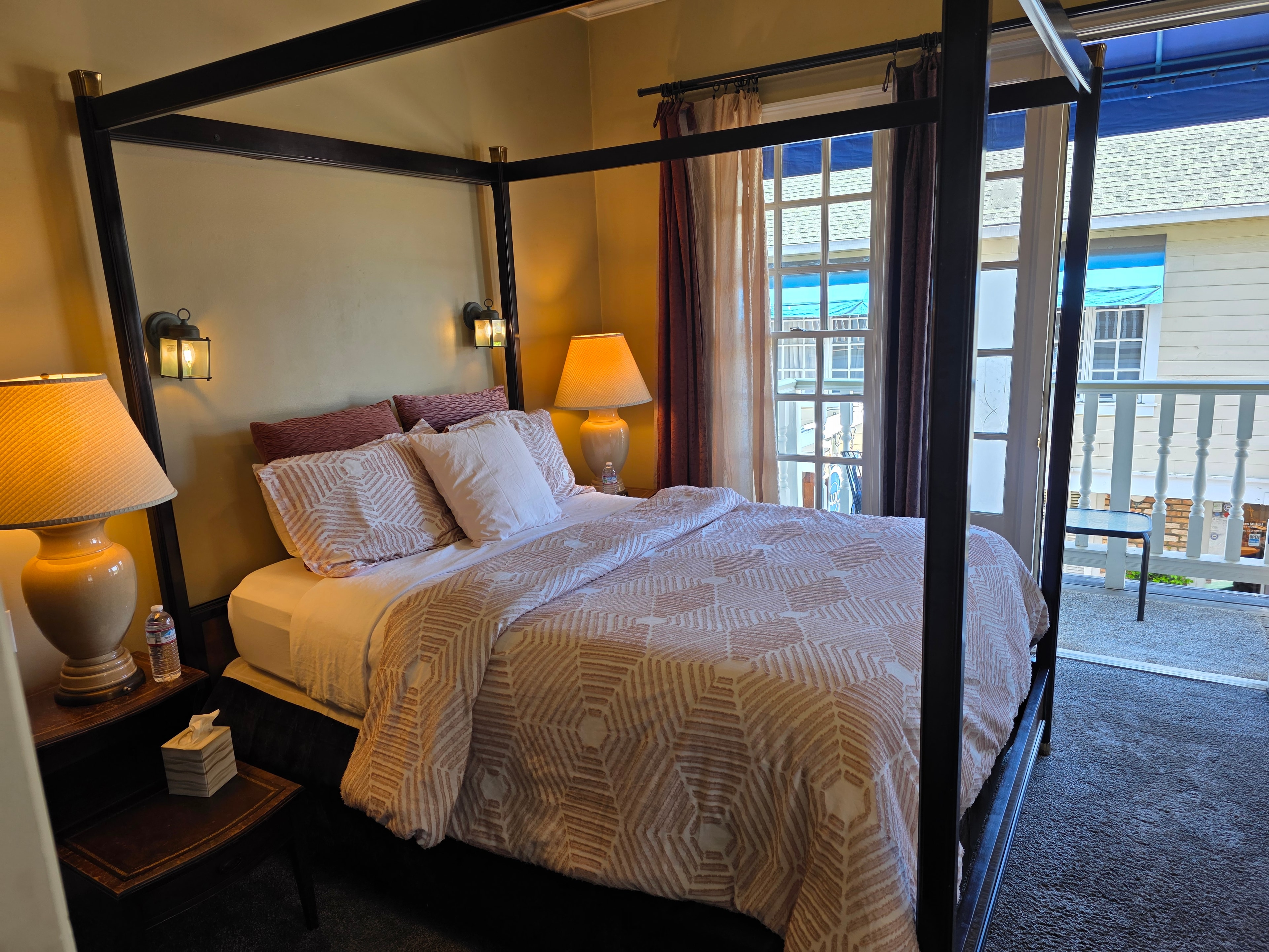 A cozy bedroom featuring a four-poster bed, warm lighting, and a view through open doors.