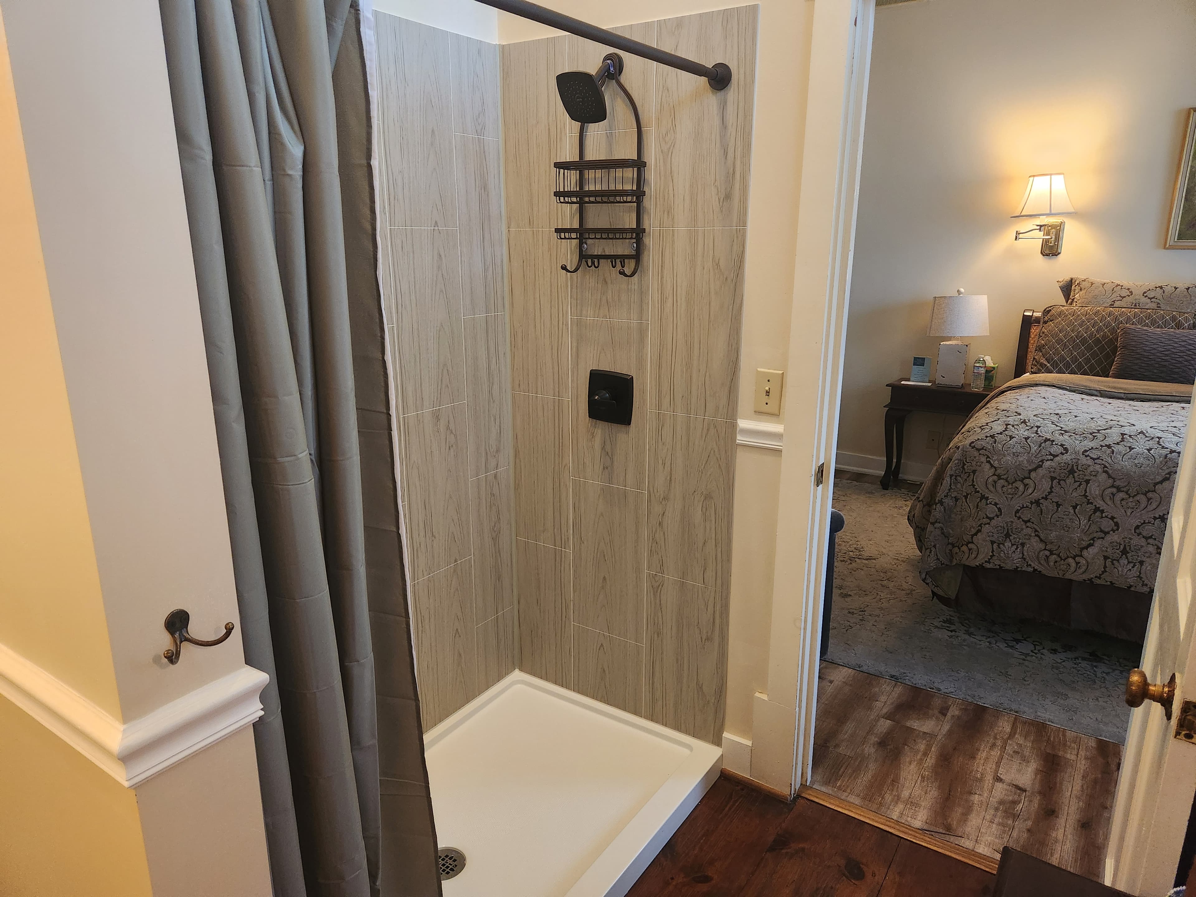 A modern shower stall with a gray curtain, leading into a bedroom with a bedside table and lamp.