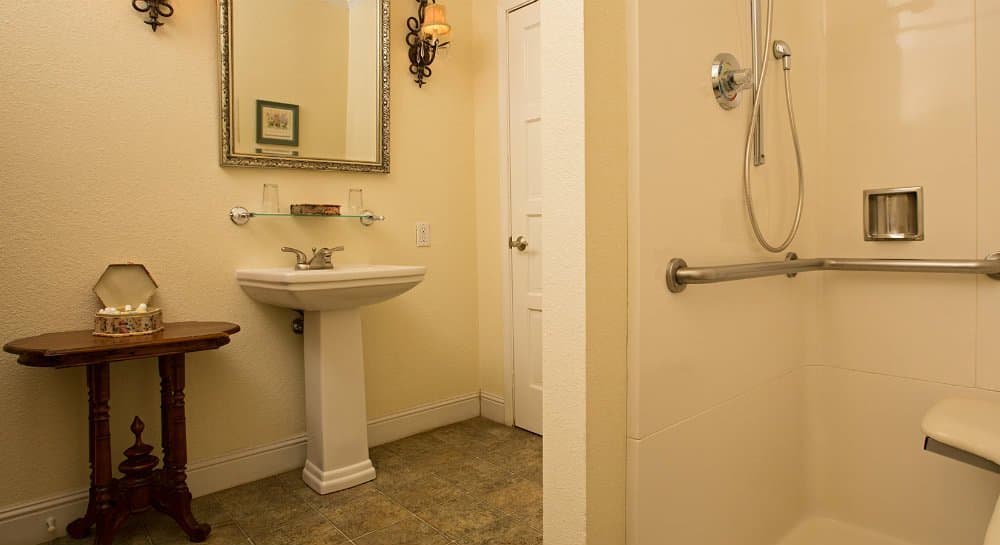 A well-lit bathroom featuring a pedestal sink, a mirror, a wooden table, and a walk-in shower.