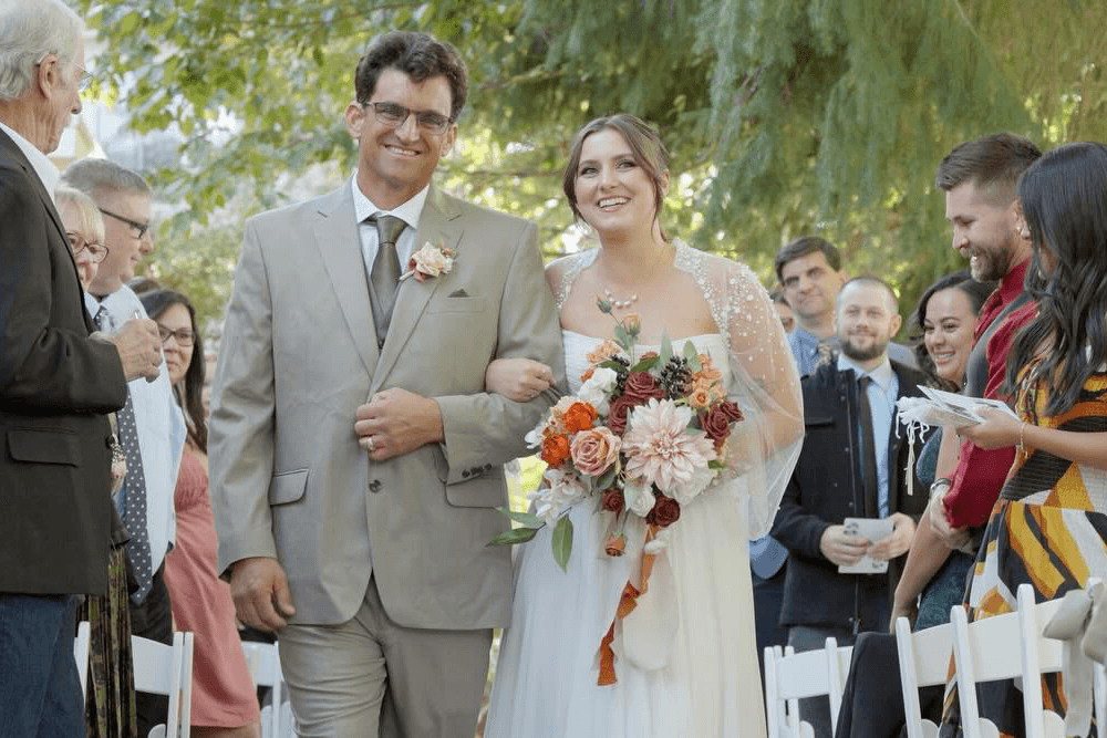 A bride walks down the aisle with her father, smiling and holding a bouquet, as guests look on.