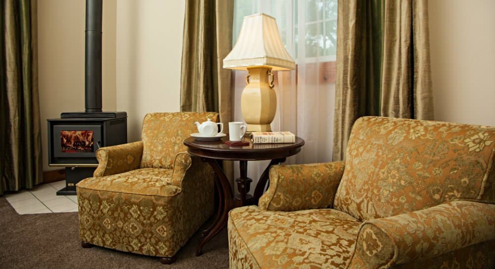 A cozy seating area with two patterned chairs, a side table holding a cup and a book, and a lamp beside a wood stove.