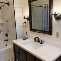A modern bathroom featuring a white sink, dark cabinetry, a mirror, and a shower area with marble tiles.