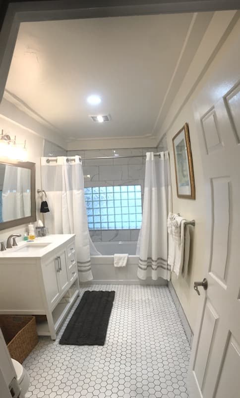 A modern bathroom featuring a white vanity, bathtub with glass block window, and hexagonal tile flooring.