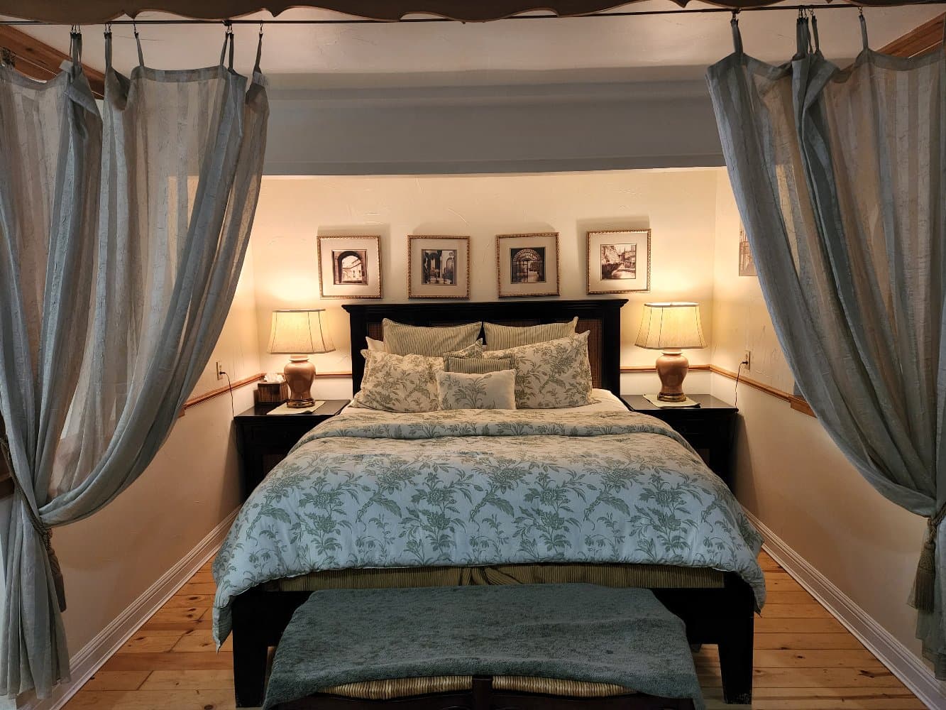 A cozy bedroom with a patterned bedspread, soft lighting from bedside lamps, and sheer curtains framing the entrance.
