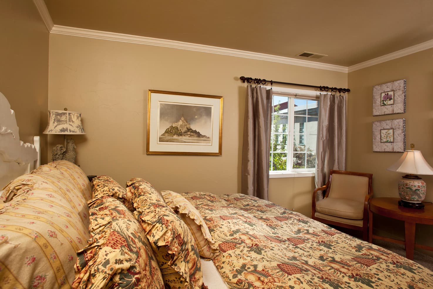 A cozy bedroom featuring a floral-patterned bedspread, framed art, and a window with curtains.