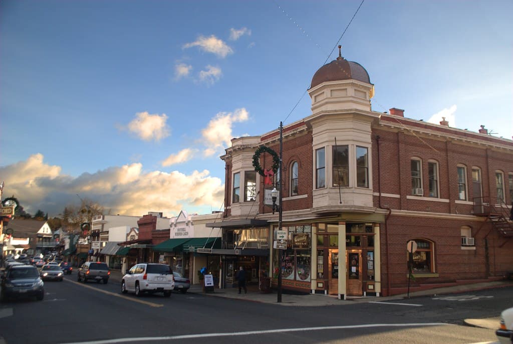 A historic brick building with a turret faces a bustling small-town street under a cloudy sky.
