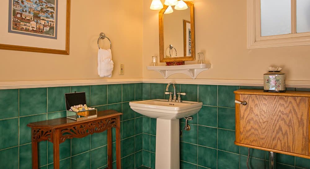 A small bathroom features a green tiled wall, a white sink, a mirror, and a wooden cabinet.
