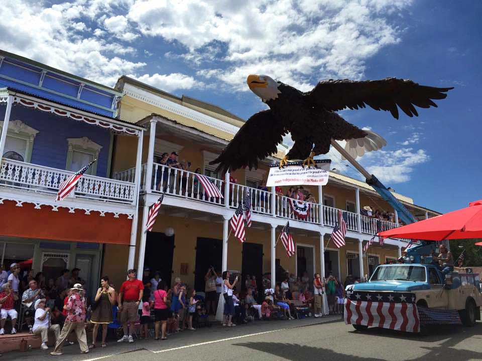 A large eagle sculpture soars above a crowd gathered in front of decorated buildings during a festive parade.