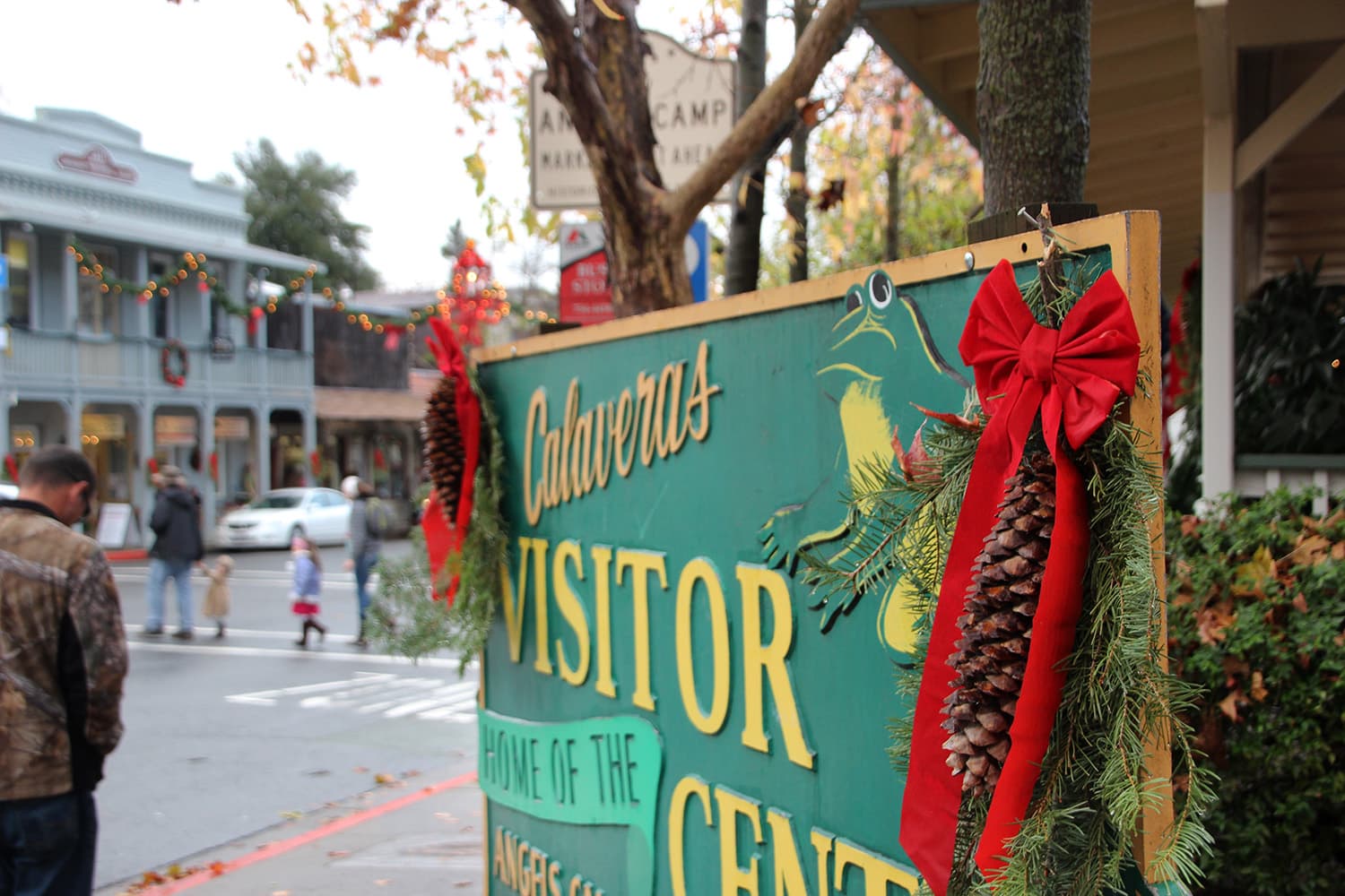 Holiday-decorated "Calaveras Visitor Center" sign with red bows, pinecones, greenery, and pedestrians on a tree-lined street. Holiday-decorated "Calaveras Visitor Center" sign with red bows, pinecones, greenery, and pedestrians on a tree-lined street.