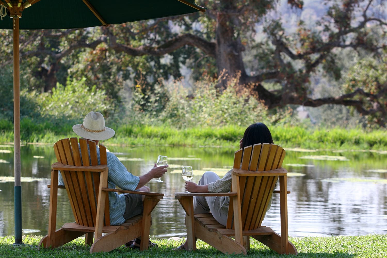 Two people relax in Adirondack chairs by a tranquil pond, holding drinks beneath an umbrella, surrounded by leafy trees and greenery. Two people relax in Adirondack chairs by a tranquil pond, holding drinks beneath an umbrella, surrounded by leafy trees and greenery.