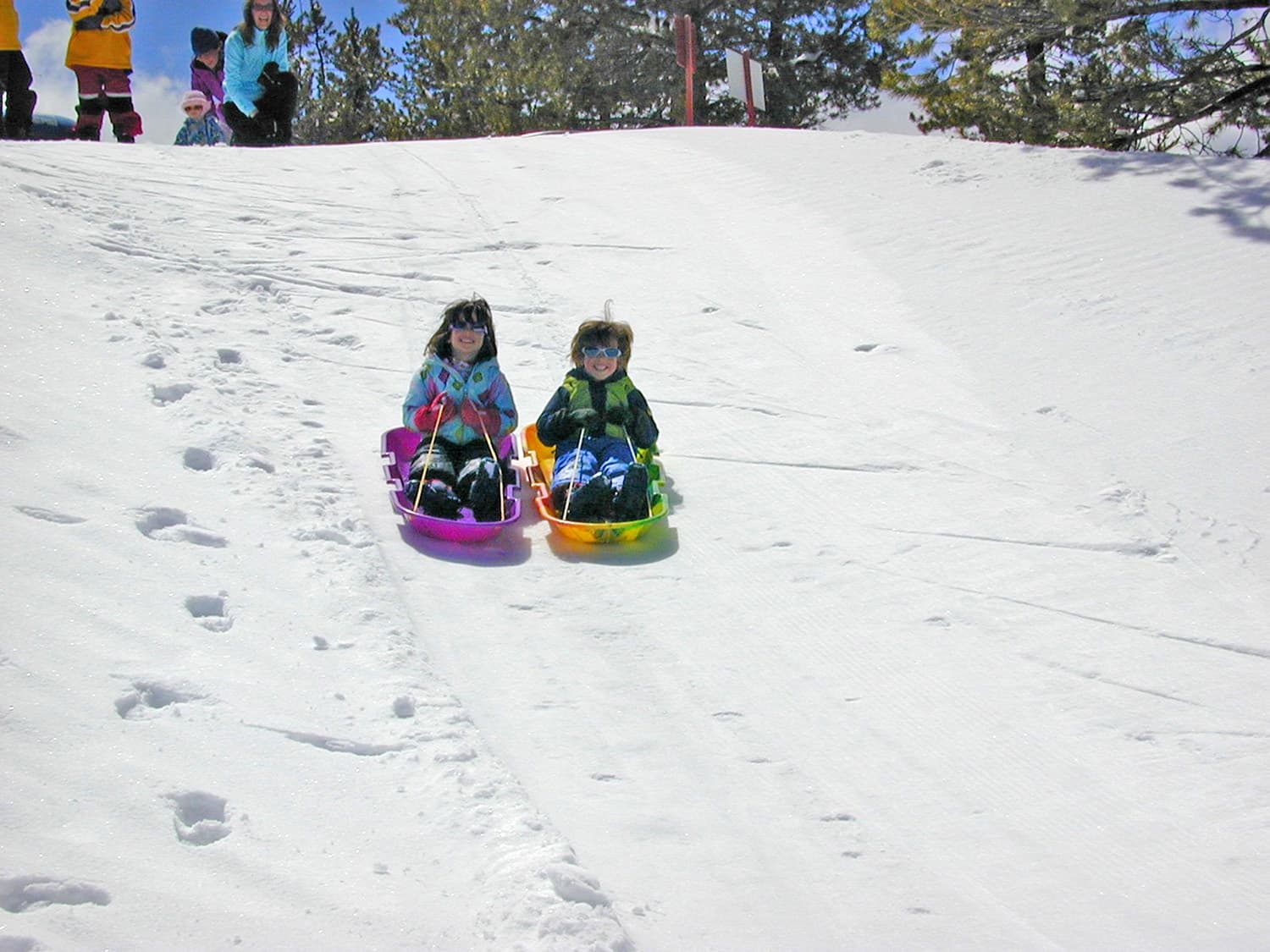 Two smiling children ride colorful sleds down a snowy hill, with footprints, evergreen trees, and onlookers above; a joyful winter scene. Two smiling children ride colorful sleds down a snowy hill, with footprints, evergreen trees, and onlookers above; a joyful winter scene.