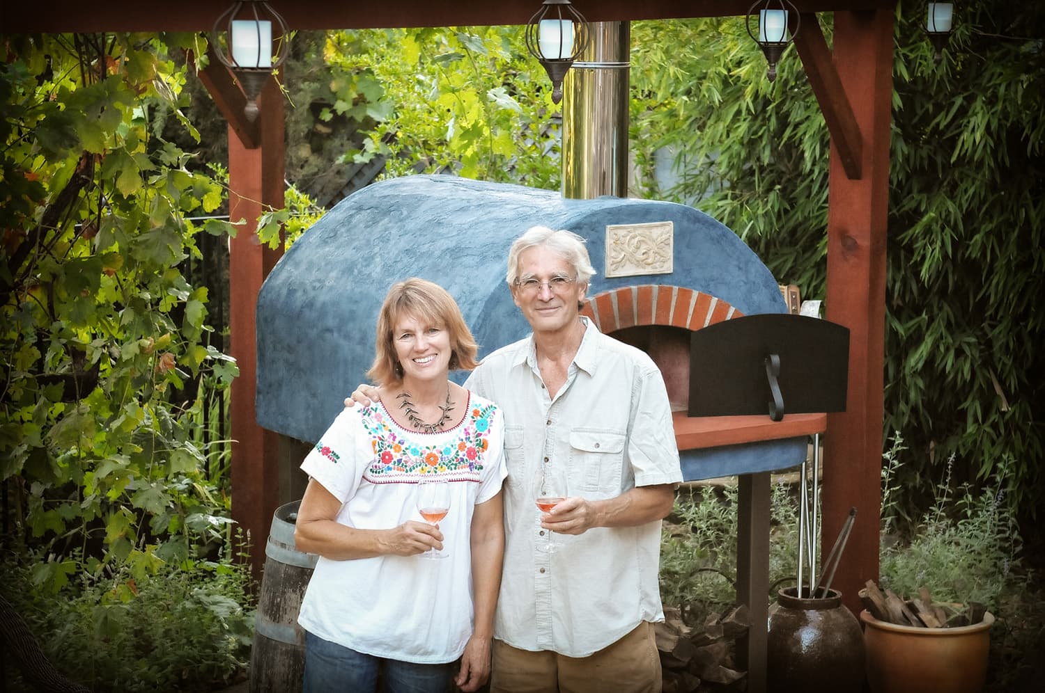 Smiling couple poses beside a rustic wood-fired pizza oven beneath a garden pergola, surrounded by lush greenery and hanging lanterns. Smiling couple poses beside a rustic wood-fired pizza oven beneath a garden pergola, surrounded by lush greenery and hanging lanterns.