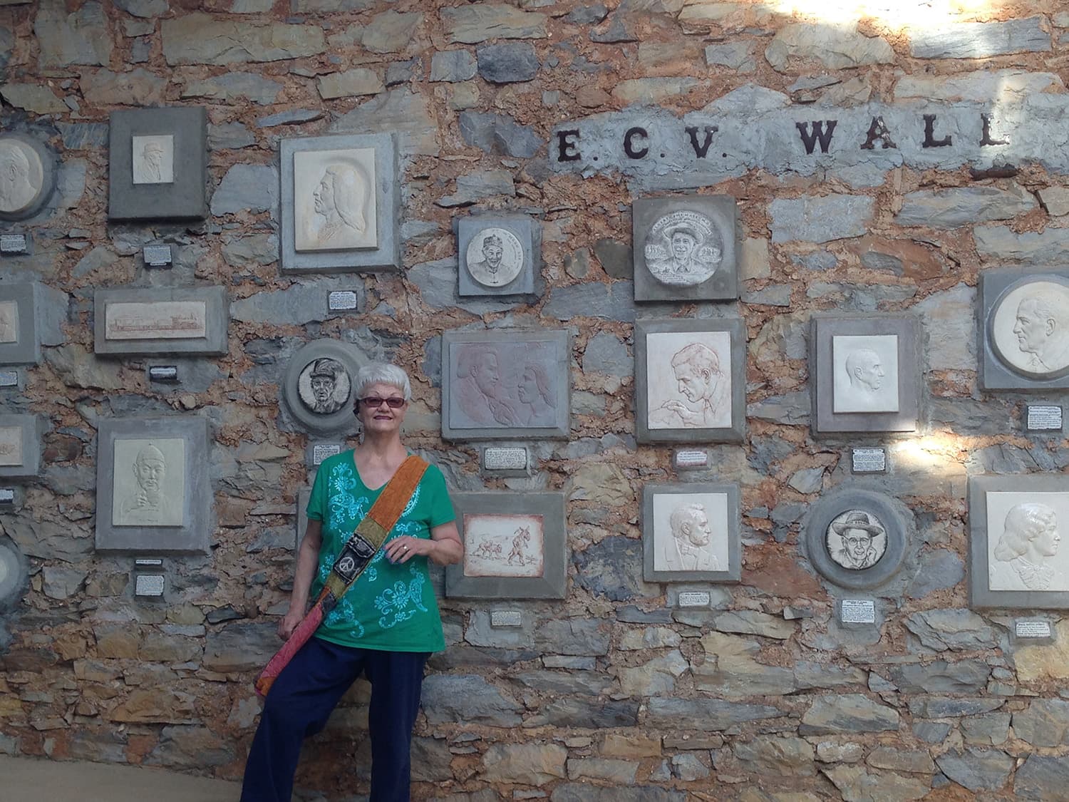 Smiling woman poses before an E.C.V. Wall, a rustic stone display covered with framed portraits, sculptures, and artwork in sunlight outdoors.