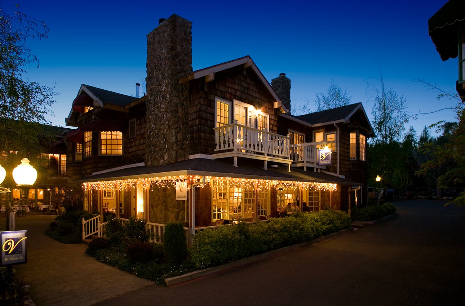Twilight view of a rustic wooden lodge glowing with string lights, a tall stone chimney, balcony, trees, and a welcoming atmosphere. Twilight view of a rustic wooden lodge glowing with string lights, a tall stone chimney, balcony, trees, and a welcoming atmosphere.