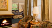 Two chairs with small table on wood floors near fireplace, and blue carpeted bedroom in background with arch top window