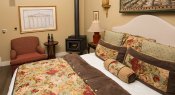 Bed with red, white, green, blue and brown floral comforter, tan walls, red chair with small table and wood stove
