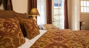 Close up of bed with rich red and gold pillows and bedspread, bedside lamp, leather chair and French doors