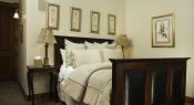 Dark wood and leather bed with ivory comforter, light green and ivory pillows, gold-framed pictures, and nightstands with lamps