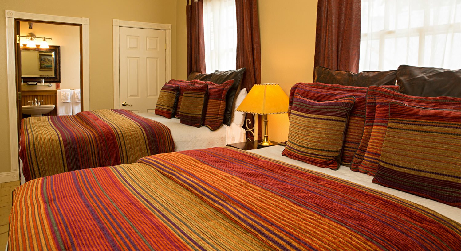 Bedroom with two beds covered in red and gold striped bedspreads, a nightstand, and two large windows