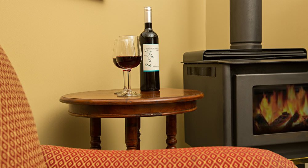 Small round wood table with wine bottle and two glasses of red wine and a black wood stove in background