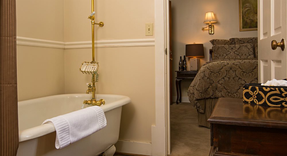 Clawfoot tub with brass faucet, ivory walls, bed in the background with brown and black comforter and dimly lit sconce