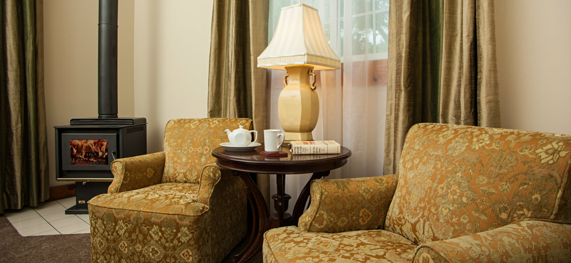 Two gold upholstered chairs flanking a small round table with ivory lamp and a black woodstove