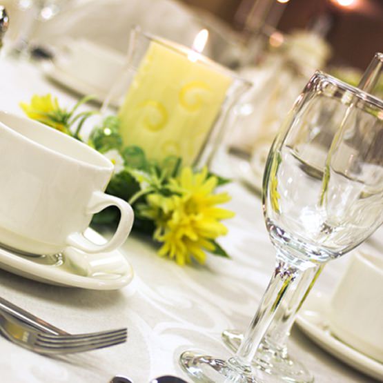 Dining table with white coffee cup and saucer, wine glasses, and a candle-lit centerpiece with greenery and yellow flowers