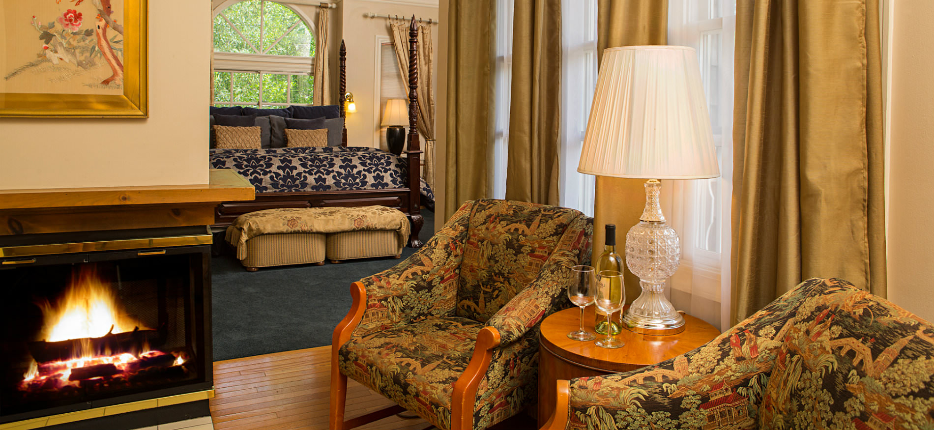 Two chairs with small table on wood floors near fireplace, and blue carpeted bedroom in background with arch top window