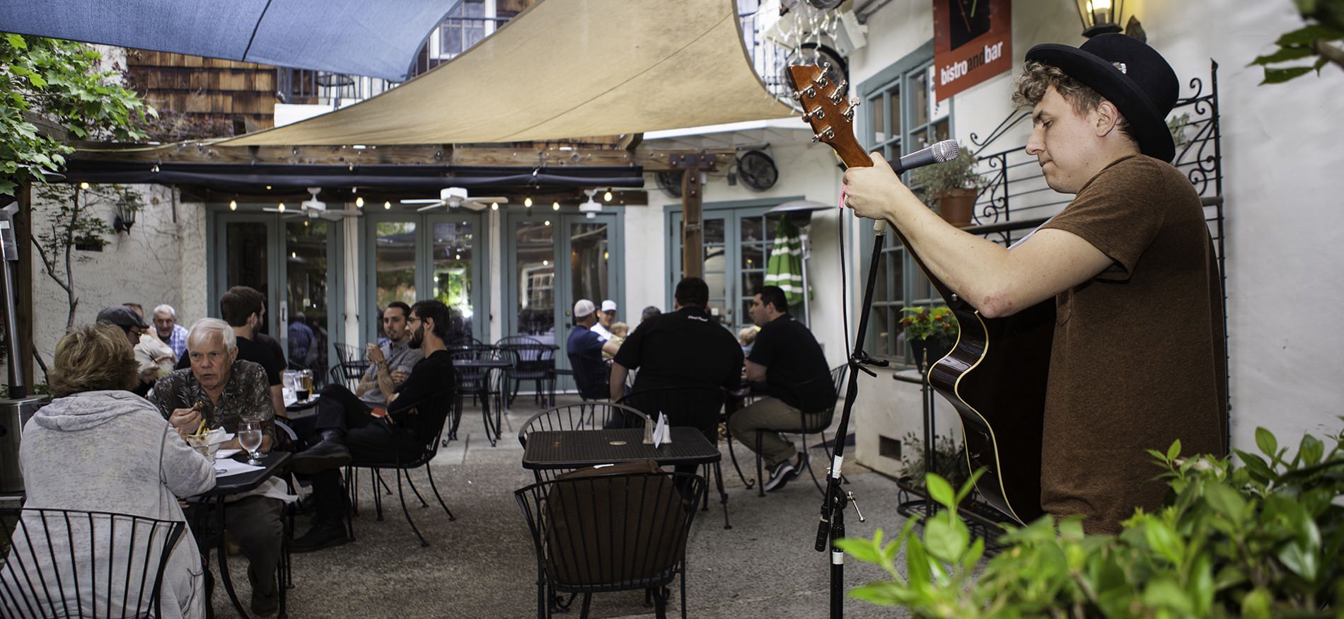 People sitting at black metal tables eating and talking outside a bistro and bar with live musician playing guitar