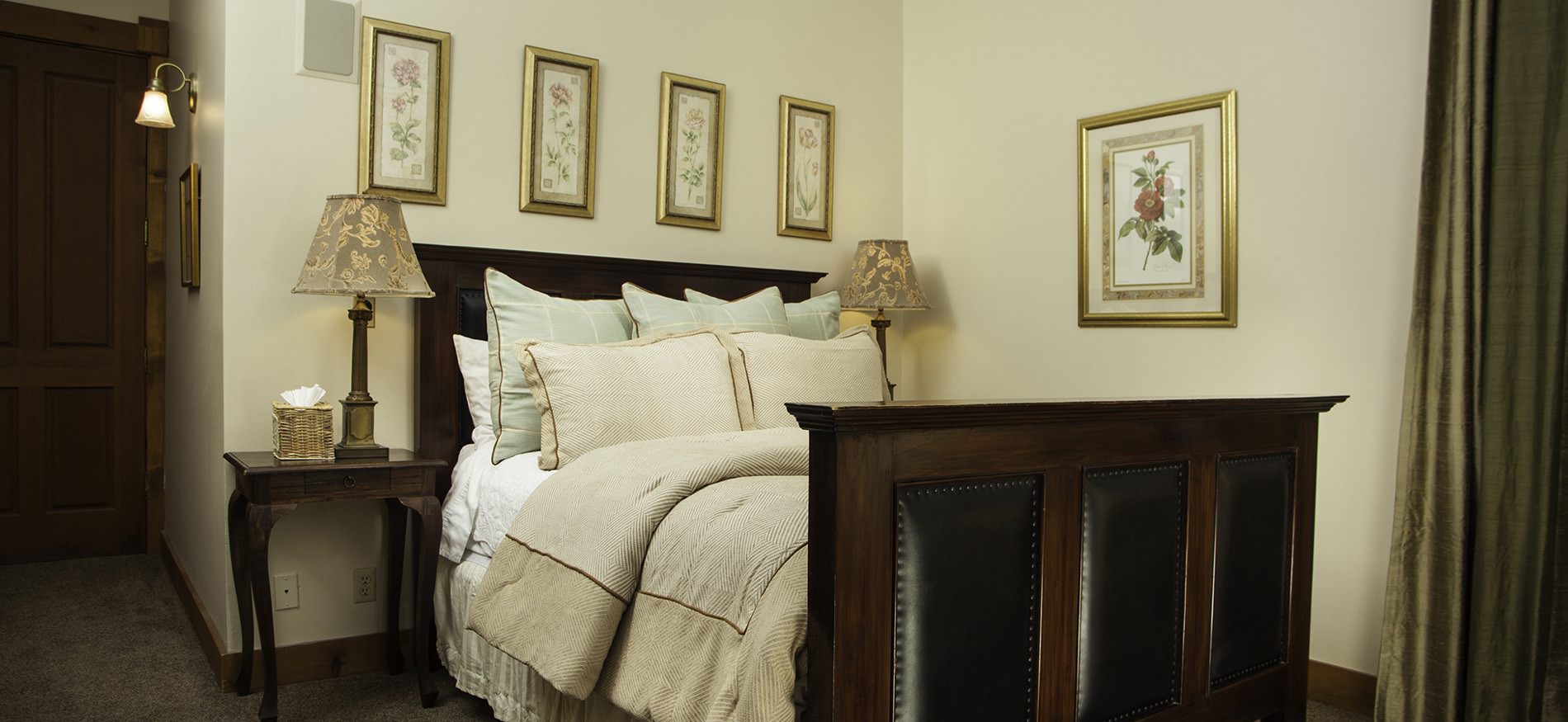 Dark wood and leather bed with ivory comforter, light green and ivory pillows, gold-framed pictures, and nightstands with lamps