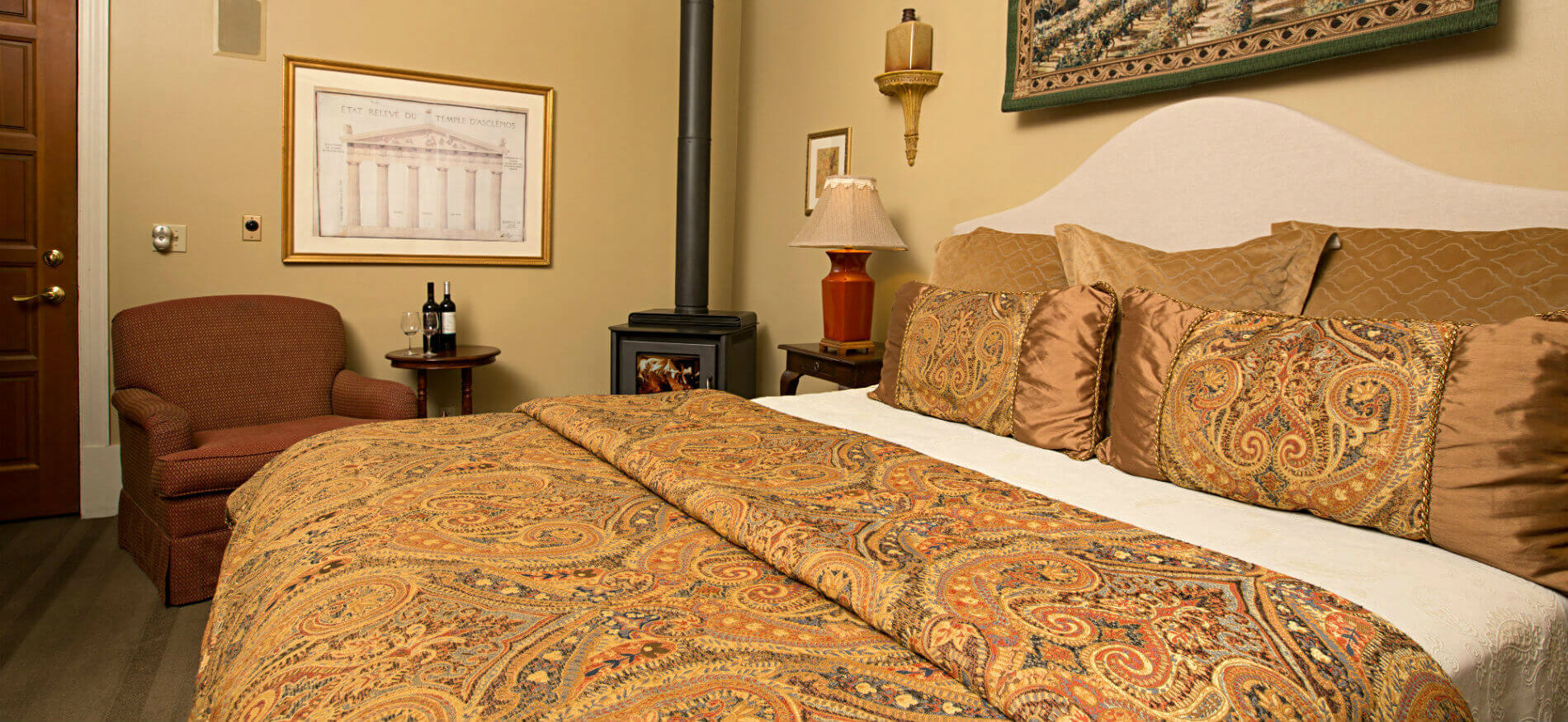 Bed with gold, blue, red, brown paisley pattern comforter, tan walls, red chair with small table, and wood stove