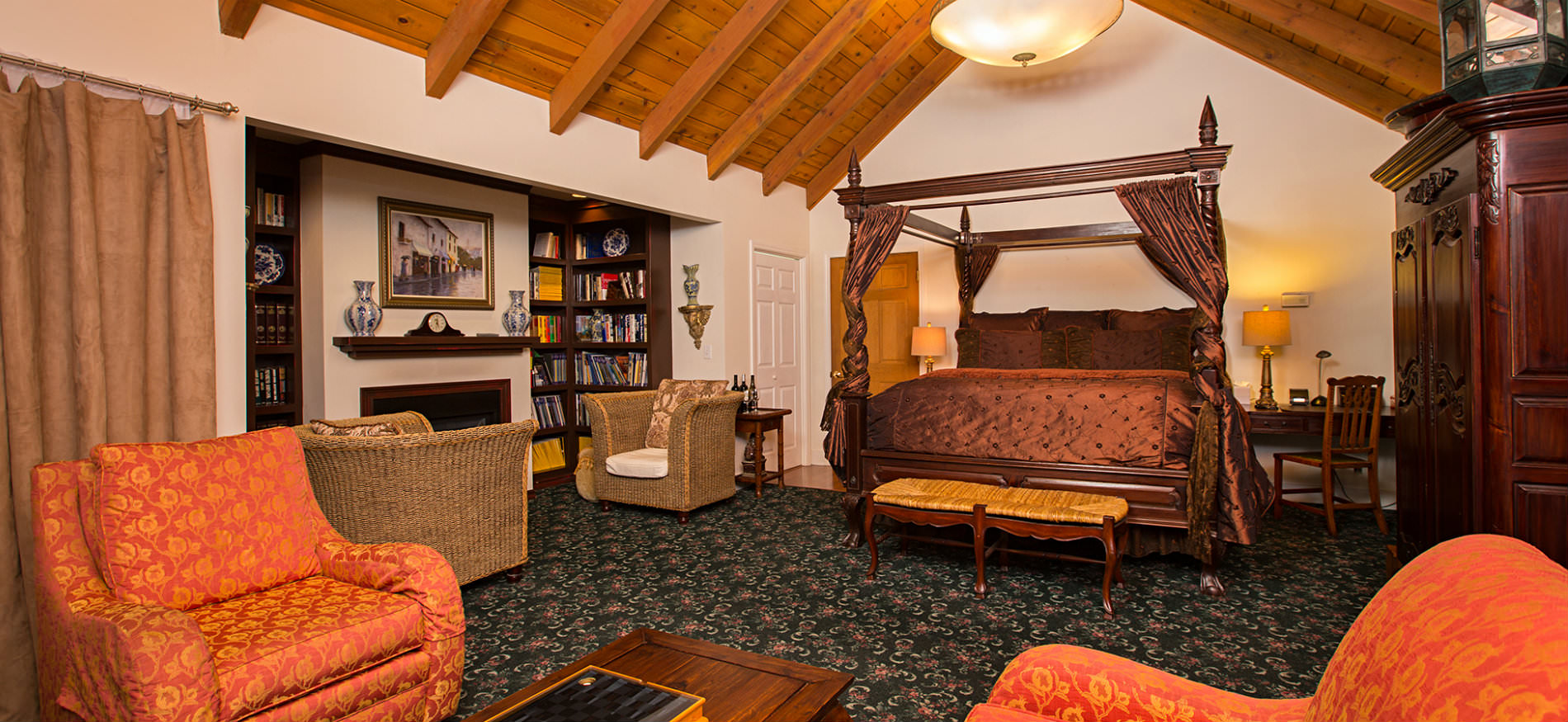 Large vaulted suite with four poster bed, bookshelves flanking fireplace with two chairs, and orange chairs with coffee table