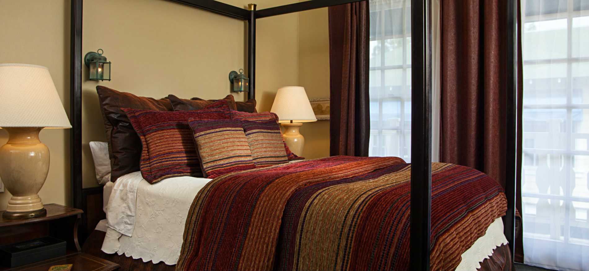 Dark four poster bed with red, brown and gold striped bedspread, large window with red curtains, two nightstands with lamps