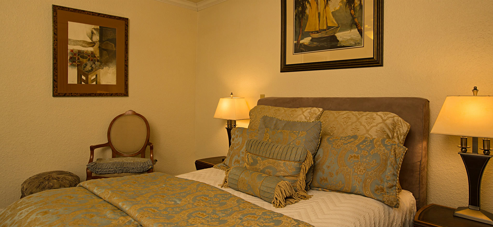 Bedroom with upholstered headboard, gold and light blue comforter, textured ivory walls, and two nightstands with lamps