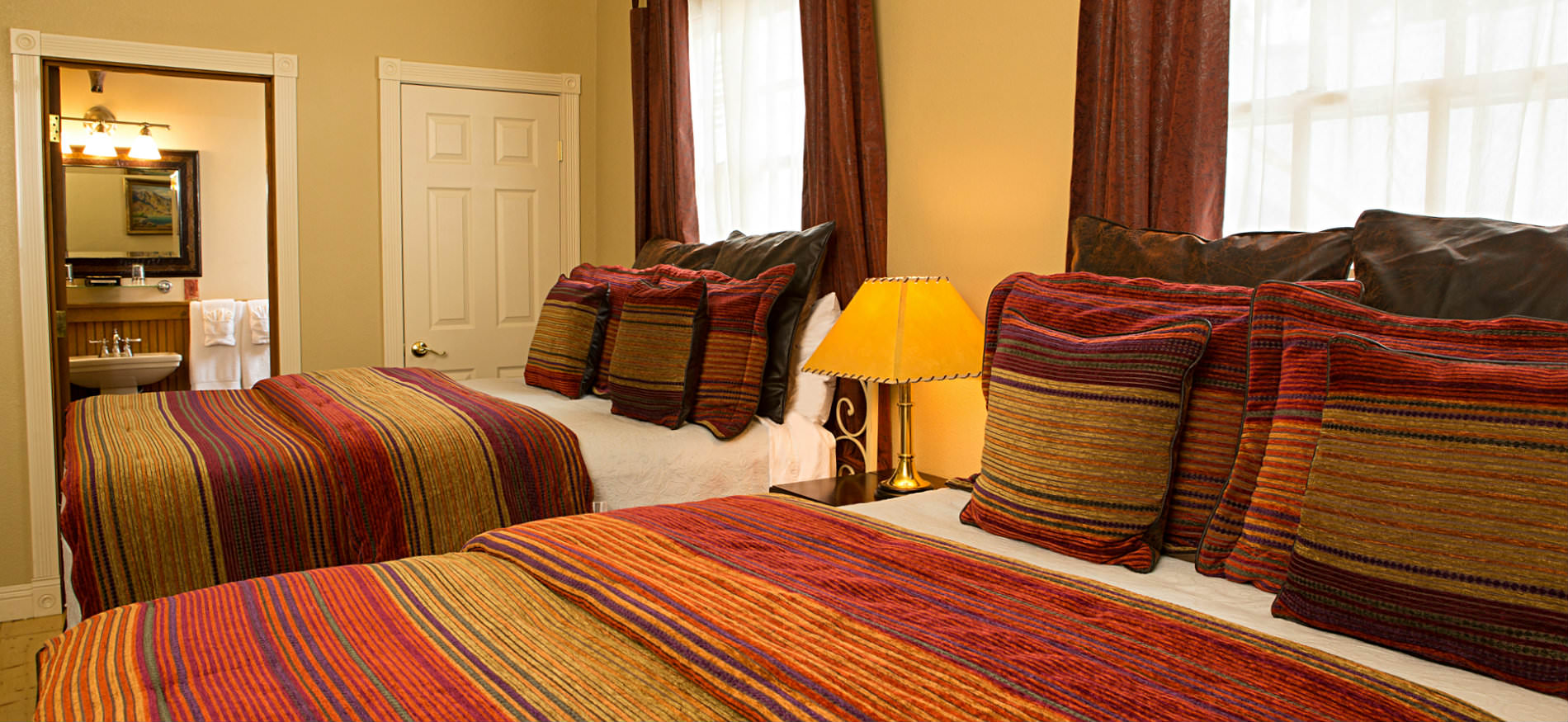 Bedroom with two beds covered in red and gold striped bedspreads, a nightstand, and two large windows