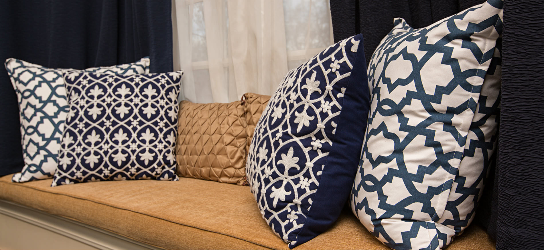 Window seat with a gold cushion and pillow and four pillows in navy and white with white sheers and navy curtains