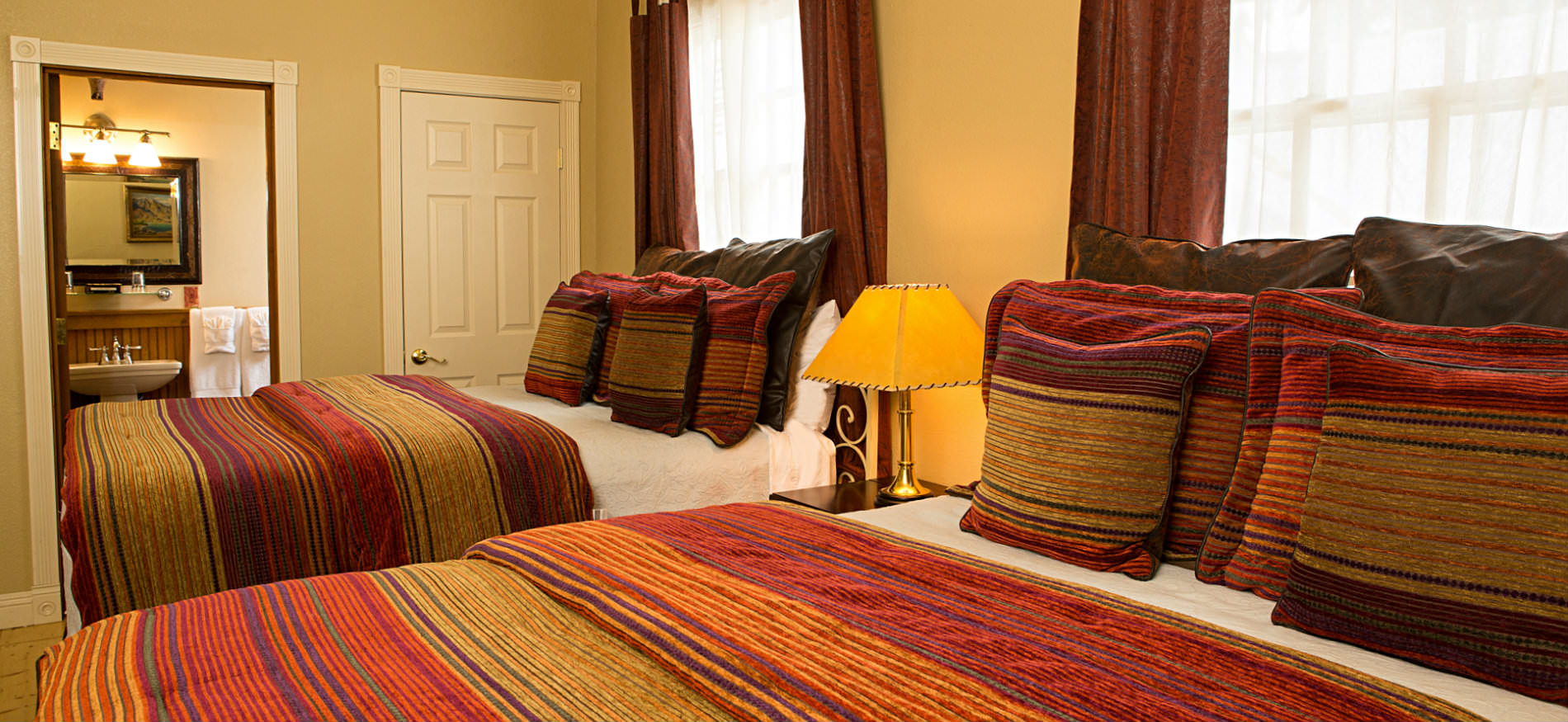 Bedroom with two beds covered in red and gold striped bedspreads, a nightstand, and two large windows
