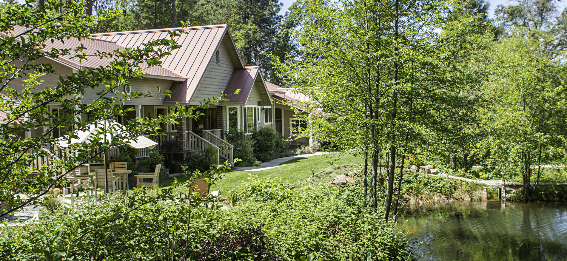 Exterior view of tan cottage with red metal gable roof, lots of lush greenery, a pond and blue skies