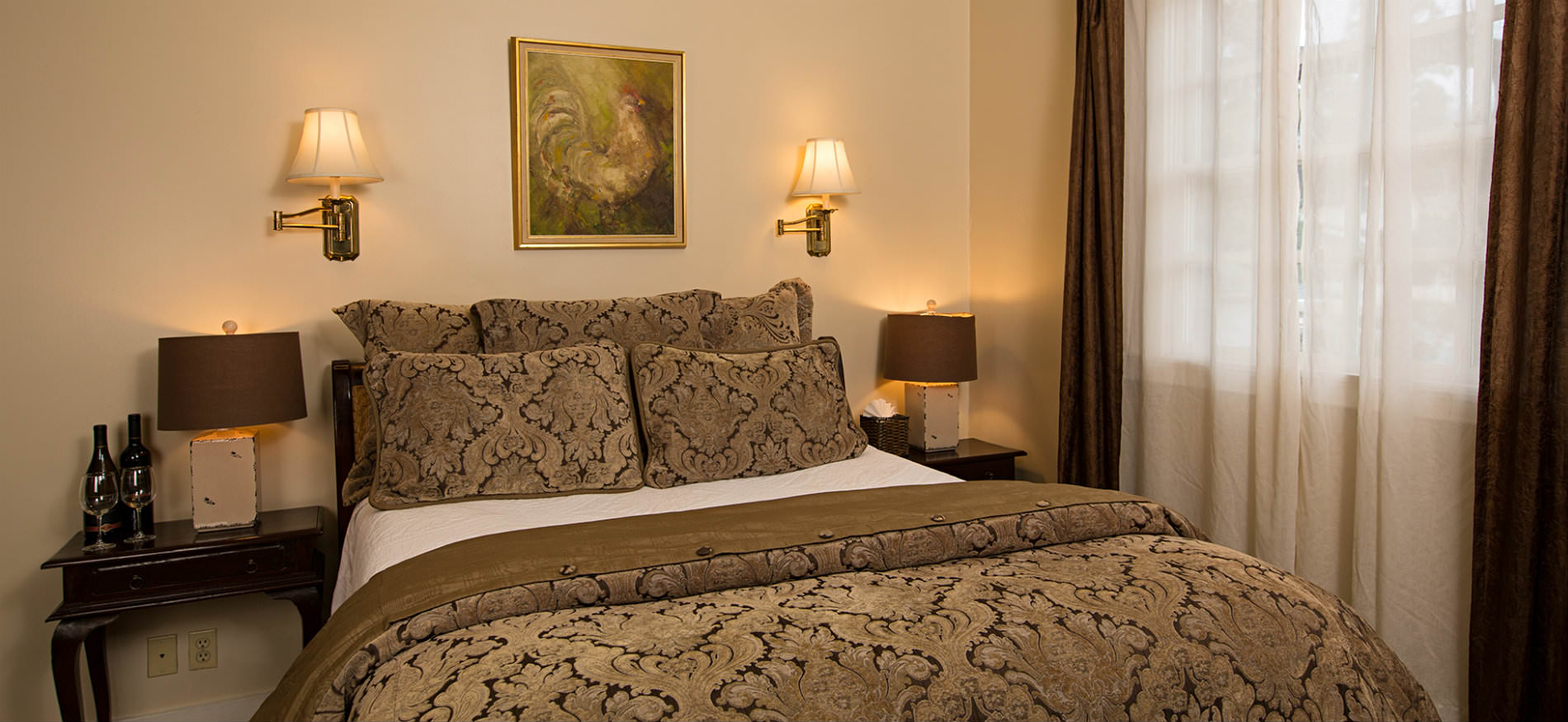 Ivory bedroom, brown pillows and comforter on bed flanked by nightstands with lamps, window with white sheers and brown curtains