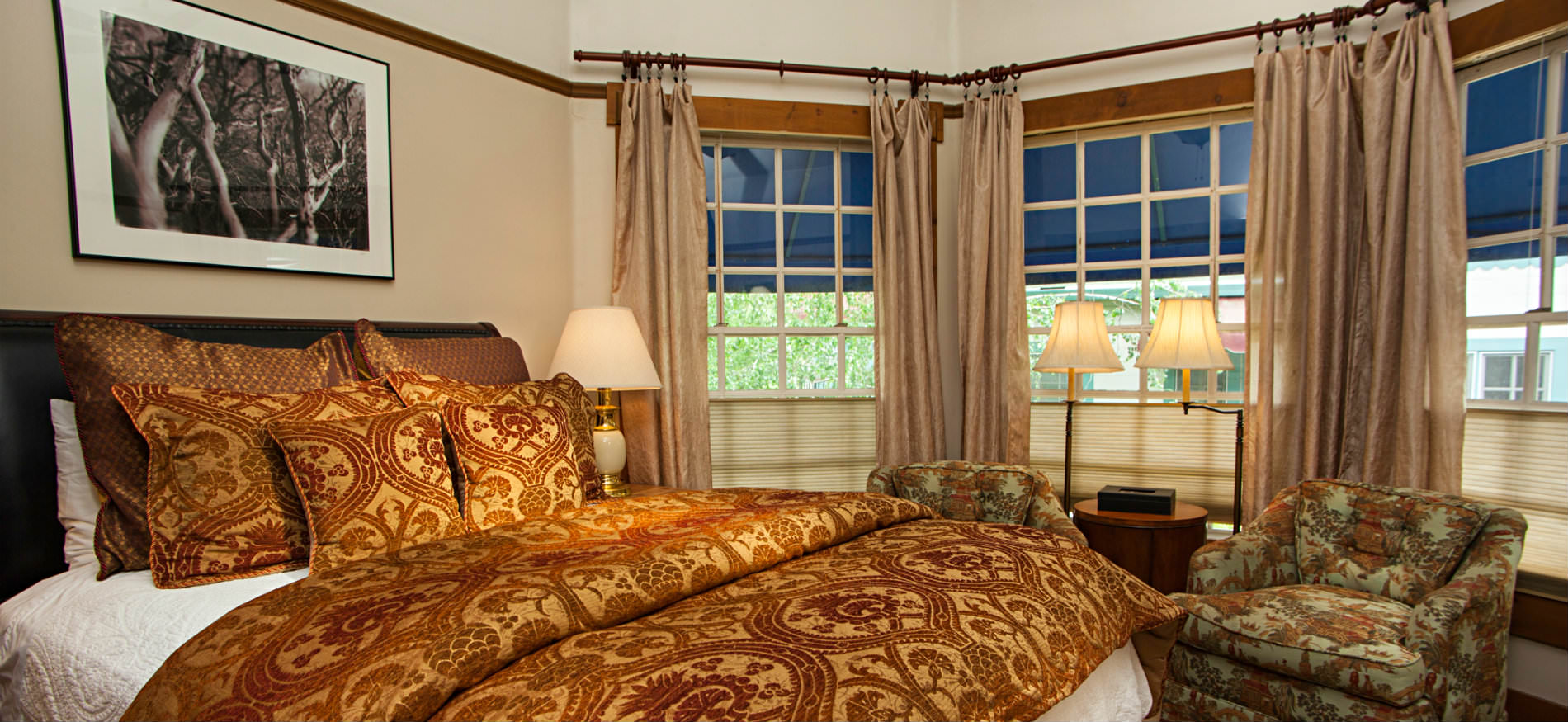 Bedroom with bay window, bed covered in red and gold pillows and comforter, two chairs and small table